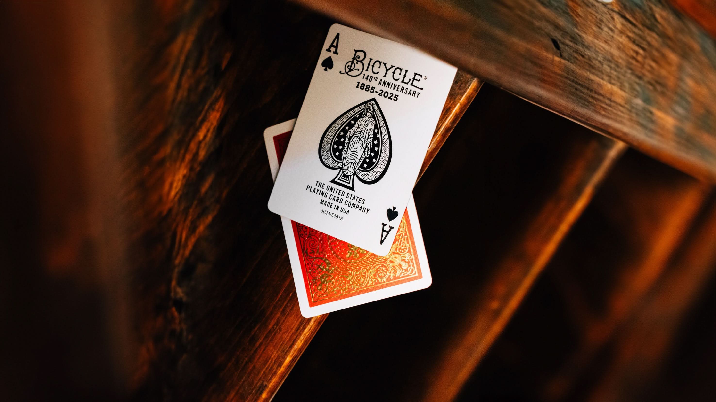 full wide shot of Bicycle 140th Anniversary playing cards ace and back with luxurious gold foil detailing on a deep red wooden stair background. Available at 52Kards for collectors, magicians, and card enthusiasts.